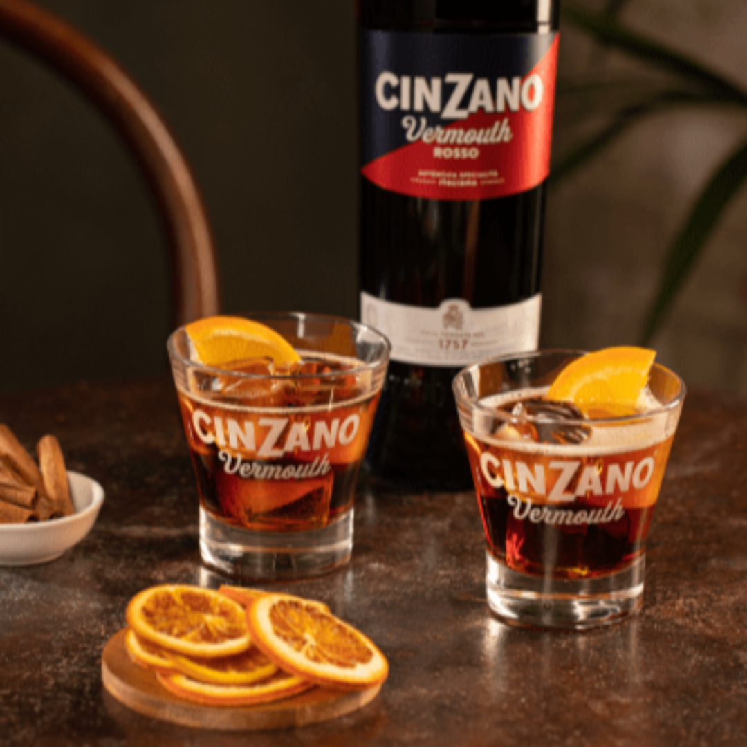 Two glasses of Cinzano Vermouth with garnishes on a dark surface, accompanied by a bottle of Cinzano Vermouth.