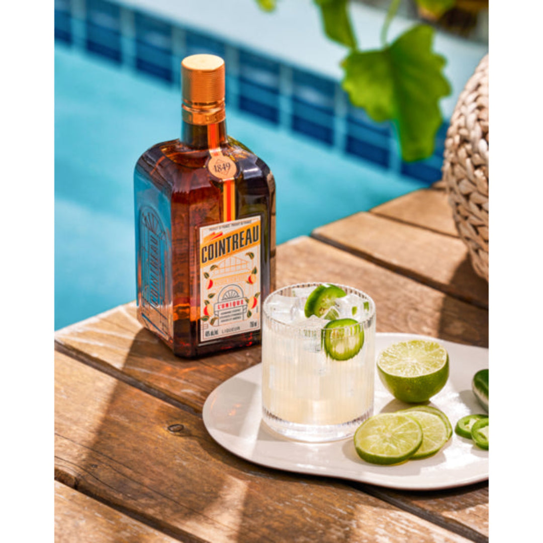 Cointreau bottle with a cocktail and lime slices on a wooden table by a pool.