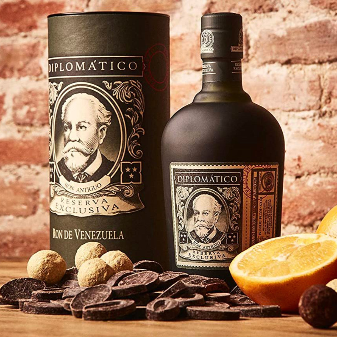 Diplomatico rum bottle and packaging with oranges and chocolates on a wooden surface against a brick wall.