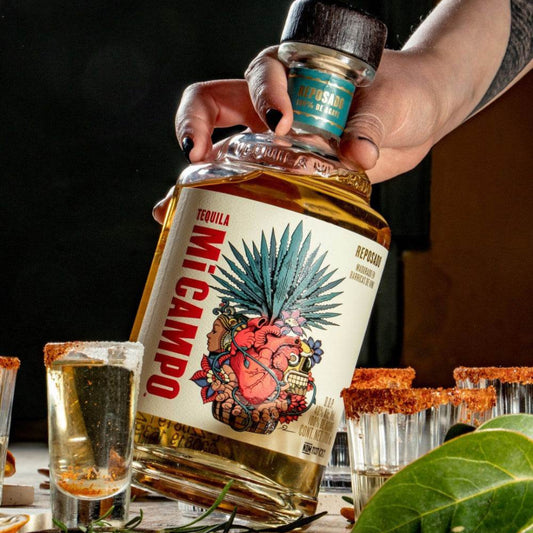Hand holding a bottle of Mi Campo tequila with shot glasses and a leaf on a wooden surface.