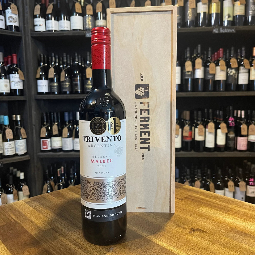 Trivento wine bottle with wooden box on a wooden surface, with shelves of wine bottles in the background.