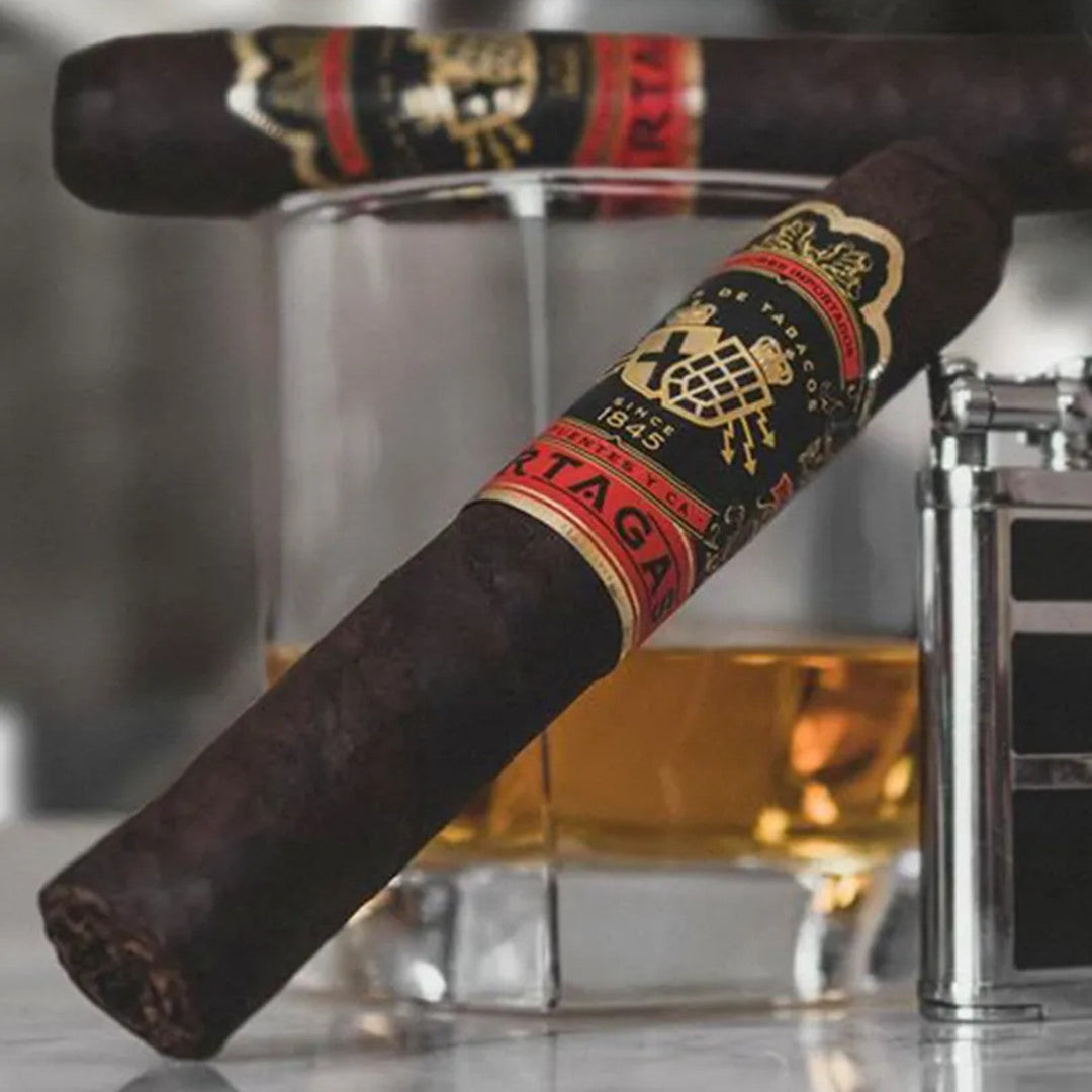 Two cigars with a prominent brand label on a reflective surface, with a glass of amber liquid in the background.