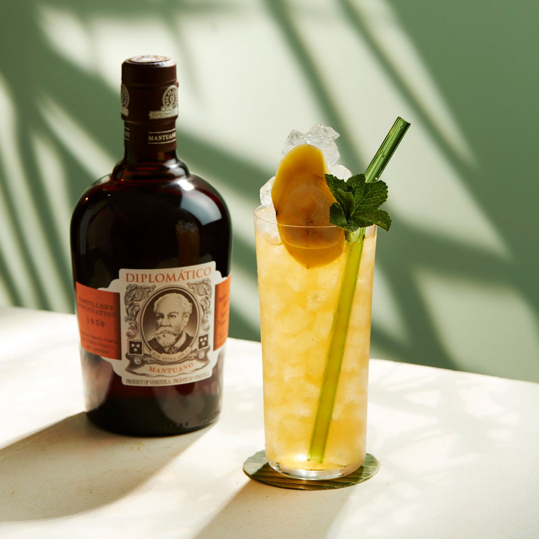 Bottle of Diplomatico rum with a cocktail garnished with lemon and mint on a white surface.