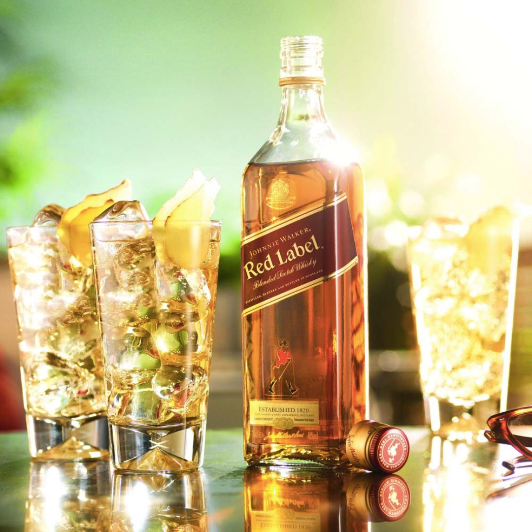 Johnnie Walker Red Label whisky bottle with glasses of ice and lemon slices on a reflective surface.