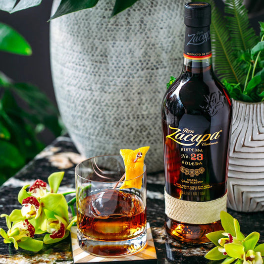 Bottle of Ron Zacapa rum with a glass of rum on a table with plants in the background